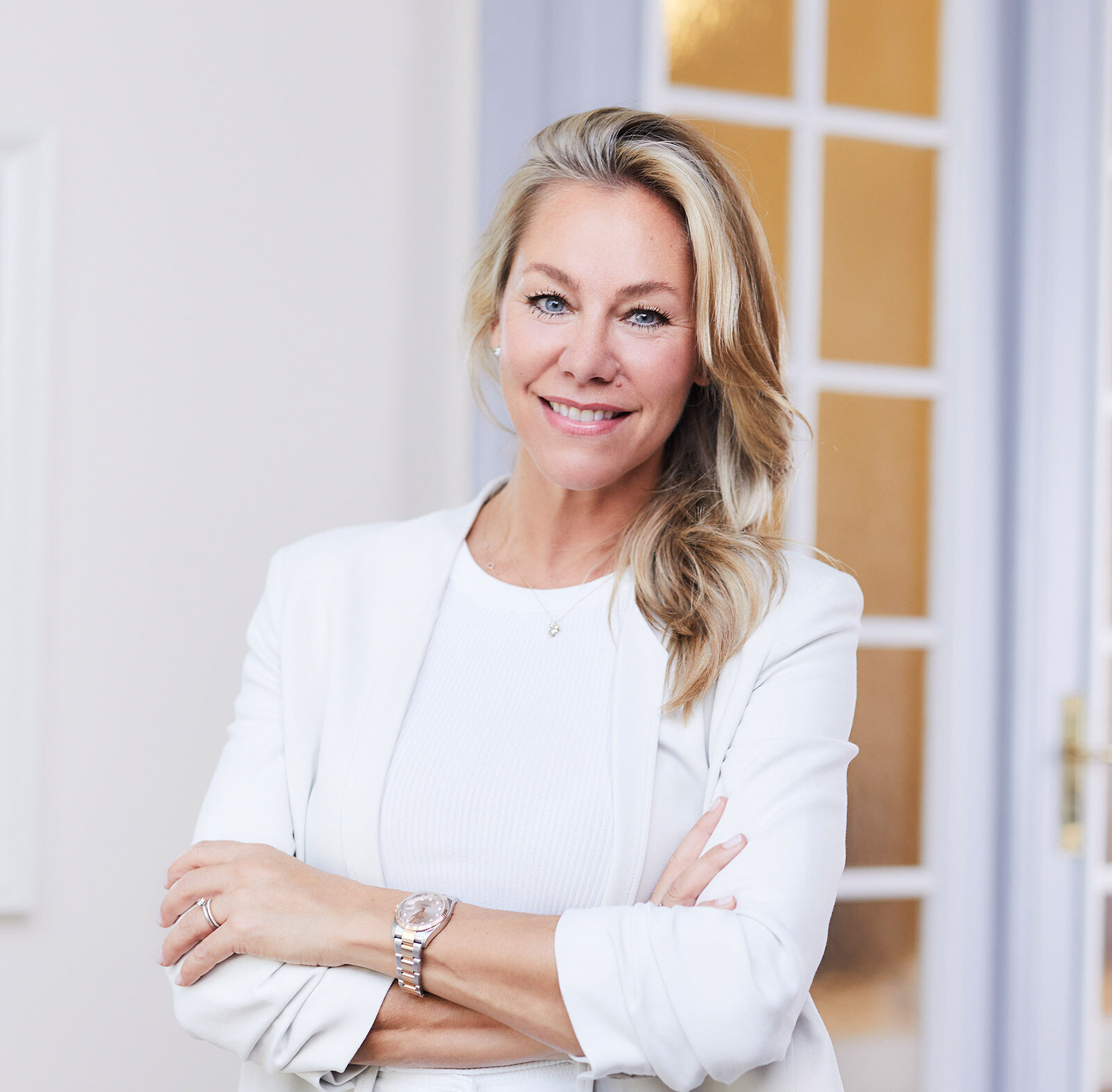 Blonde Frau mit freundlichem Lächeln trägt weißen Blazer und steht vor Glastür