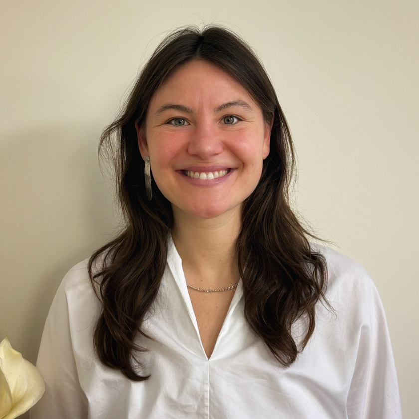 Eine junge Frau mit langen braunen Haaren lacht freundlich vor hellem Hintergrund